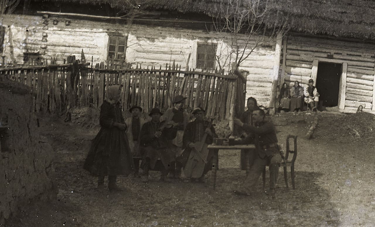 Na archiwalnym zdjęciu grupa ludzi przy stole ustawionym przed wiejską chatą