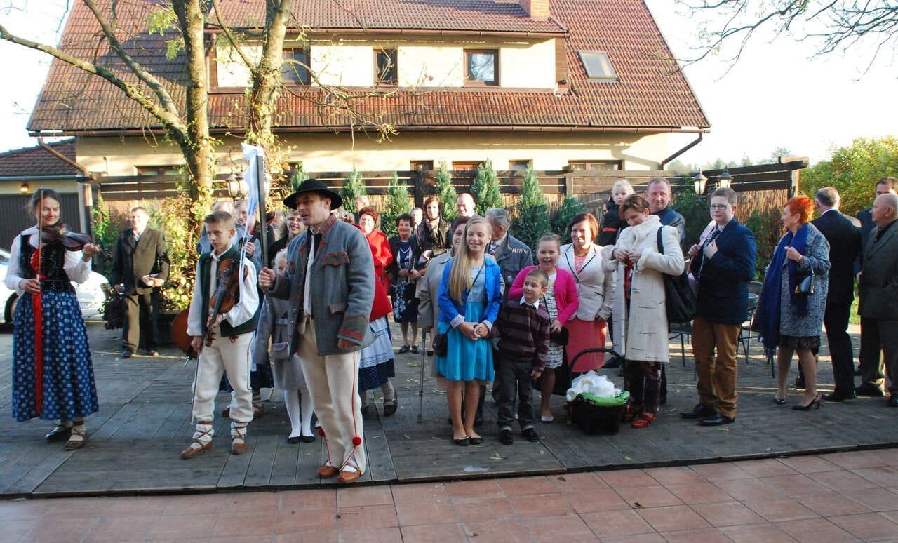 Grupa gości weselnych z drużbą i kapelą. Ludzie w różnym wieku, ubrani odświętnie